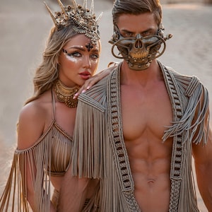 May include: A couple wearing elaborate, gold-toned, fringe outfits. The woman is wearing a crown and the man is wearing a skull-shaped mask. They are standing in a desert setting.