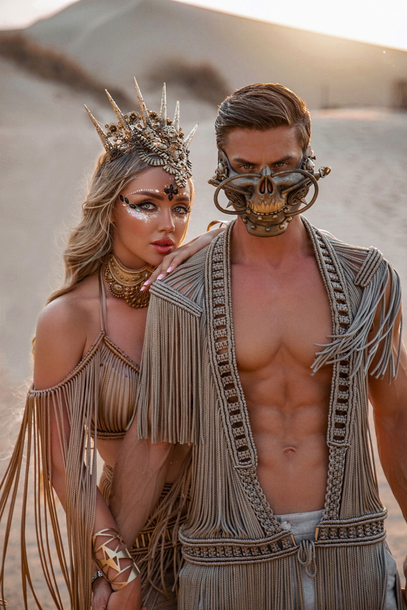 May include: A couple wearing elaborate, gold-toned, fringe outfits. The woman is wearing a crown and the man is wearing a skull-shaped mask. They are standing in a desert setting.