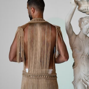 May include: A tan crocheted vest with fringe detailing. The vest is sleeveless and has a high neckline. The vest is made of a thick, textured yarn.