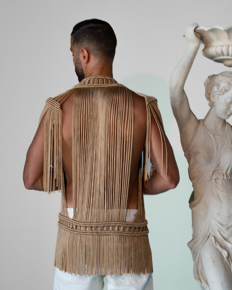 May include: A tan crocheted vest with fringe detailing. The vest is sleeveless and has a high neckline. The vest is made of a thick, textured yarn.