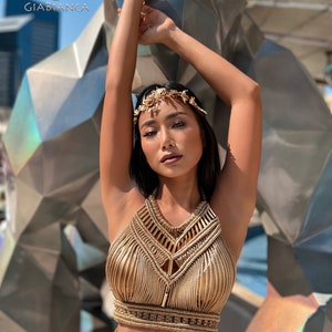 May include: A woman wearing a gold halter top and skirt made of braided rope. She is wearing a gold leaf headpiece and has her arms raised above her head.