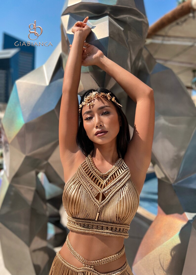 May include: A woman wearing a gold halter top and skirt made of braided rope. She is wearing a gold leaf headpiece and has her arms raised above her head.
