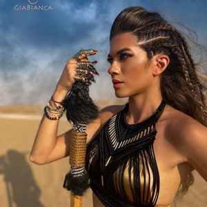 May include: A woman with long brown hair styled in braids wears a black and gold halter top with a geometric design. She holds a wooden staff with a black and white patterned wrap and a black fur pom-pom.