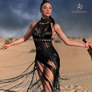 K&ouml;nnte beinhalten: Eine Frau in einem schwarzen Fransenkleid mit goldenen Akzenten steht in einer W&uuml;stenlandschaft. Sie h&auml;lt eine Geige. Der Text "Giabianca" ist in der oberen rechten Ecke des Bildes sichtbar.