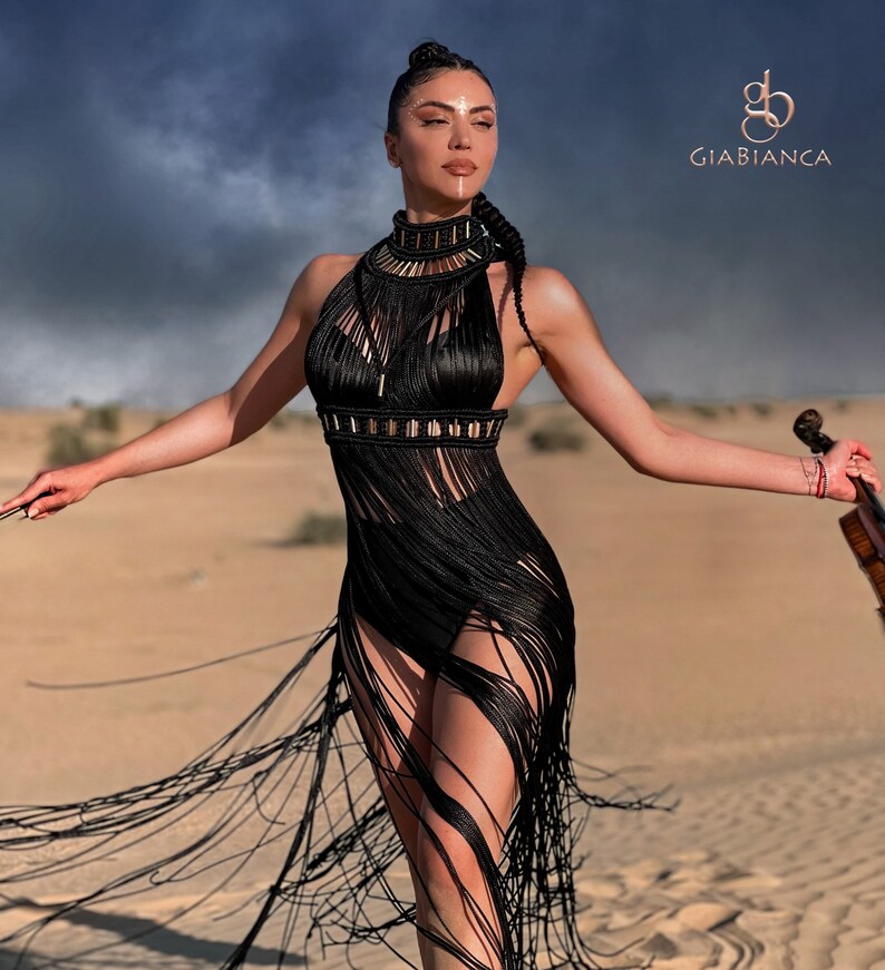 K&ouml;nnte beinhalten: Eine Frau in einem schwarzen Fransenkleid mit goldenen Akzenten steht in einer W&uuml;stenlandschaft. Sie h&auml;lt eine Geige. Der Text "Giabianca" ist in der oberen rechten Ecke des Bildes sichtbar.