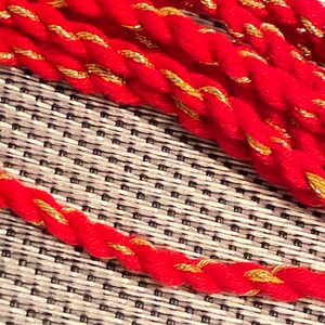 May include: Close-up of several strands of red rope with gold metallic thread woven throughout. The rope is thick and appears to be made of a soft material. The background is a textured grey and white surface.