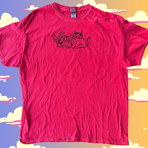 May include: A bright pink t-shirt featuring a black graphic of an angel and a devil. The angel has a halo and wings, while the devil has horns and a tail. The shirt is made of a soft material.