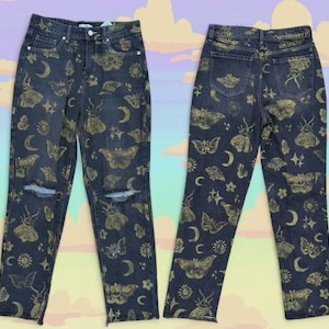 May include: Dark wash denim jeans featuring a repeating pattern of gold-colored moths, moons, and stars. The jeans have a distressed knee and a classic five-pocket design. The back of the jeans also displays the same celestial pattern.