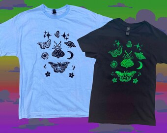 May include: Two t-shirts, one light blue and one black, each featuring a graphic design of moths, butterflies, stars, and a crescent moon. The light blue shirt has a blue collar. The black shirt has a neon green design.