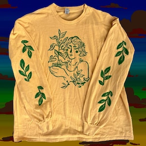 Puede incluir: Camiseta de manga larga de color amarillo mostaza con una ilustración verde de una mujer sosteniendo una planta. Diseños de hojas verdes impresos en las mangas. La camiseta es de material suave, ideal para uso diario.