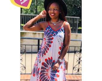 May include: A white, red, and blue patterned maxi dress with a V-neck and spaghetti straps. The dress has a high-low hemline and is made of a lightweight fabric. The model is wearing a black hat.
