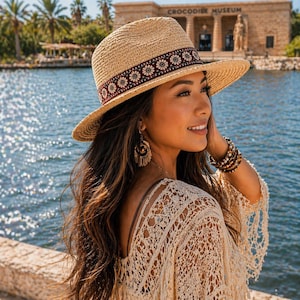 May include: A straw fedora hat with a decorative band featuring a floral pattern in red, white, and brown. The hat is worn outdoors near a body of water. The image also shows a cream-colored lace top and jewelry.