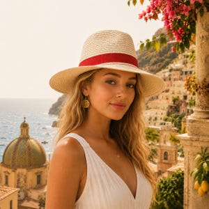 May include: A straw fedora hat with a red band. The hat has a wide brim and is worn by a person with long blonde hair. The background features a coastal town with a church dome and the sea.