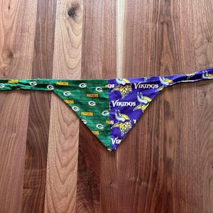 May include: A triangular pet bandana featuring a split design. The left side is green with the yellow Packers G logo and text, while the right side is purple with the Minnesota Vikings logo and text. The fabric has long ties on both ends.