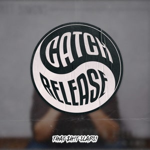 May include: Black and white yin yang symbol with the words "CATCH RELEASE" in a bold, stylized font. The text "THAT $#!T SLAPS!" is below the symbol.