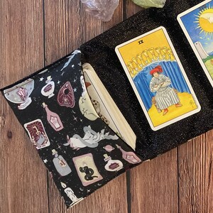 May include: A black velvet tarot card pouch with a black strap. The pouch is open, revealing three tarot cards: The Moon, The Star, and Two of Cups. The pouch is decorated with a black and white pattern of potion bottles, stars, and cats.