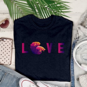 May include: A navy blue t-shirt with the word "LOVE" in gradient pink and purple letters. The "O" is replaced with a colorful betta fish design. The shirt is surrounded by a pair of blue jeans, white sneakers, a denim jacket, and headphones.