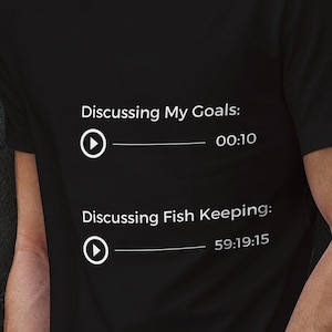 May include: Black t-shirt with white text that reads "Discussing My Goals: 00:10" and "Discussing Fish Keeping: 59:19:15". The shirt is worn by a person standing in front of a dark textured wall.