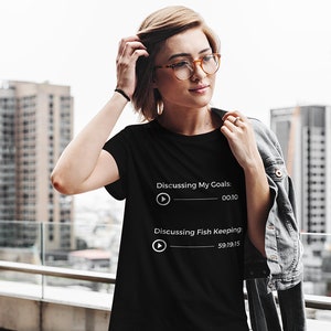 May include: Black t-shirt with white text that reads "Discussing My Goals: 00:10" and "Discussing Fish Keeping: 59:19:15". The shirt is worn by a person with glasses and a denim jacket, standing in front of a city skyline.
