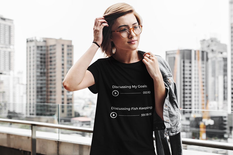 May include: Black t-shirt with white text that reads "Discussing My Goals: 00:10" and "Discussing Fish Keeping: 59:19:15". The shirt is worn by a person with glasses and a denim jacket, standing in front of a city skyline.