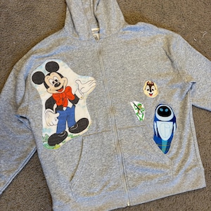 Puede incluir: Una sudadera gris con cremallera con parches de Mickey Mouse, Chip y Wall-E. El parche de Mickey Mouse es una ilustración colorida. La sudadera tiene un bolsillo delantero y una capucha.