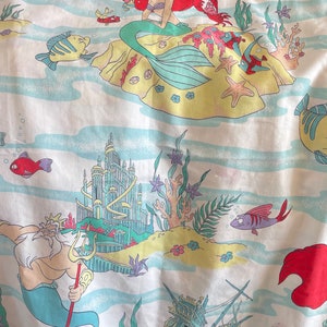May include: White fabric featuring a colorful underwater scene with Ariel the mermaid, fish, and a sunken ship. The design includes light blue waves, yellow sand, and various sea creatures. The Disney logo is visible in the top left corner.