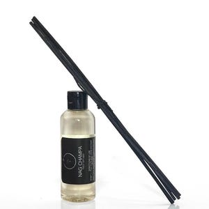 May include: A clear glass bottle with a black cap and a black label that reads "NAG CHAMPA". The bottle is filled with a light-colored liquid. A bundle of black reed diffuser sticks leans against the bottle, creating a minimalist aesthetic.