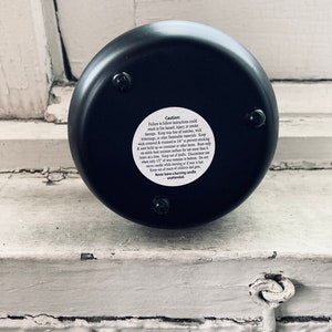 A black soy wax candle labeled “Cold Dead Heart” in gothic-style typography. It comes in a dark container with a matching lid featuring a minimalist triangle logo. The candle has a single white wick and a moody design perfect for gothic aesthetics.