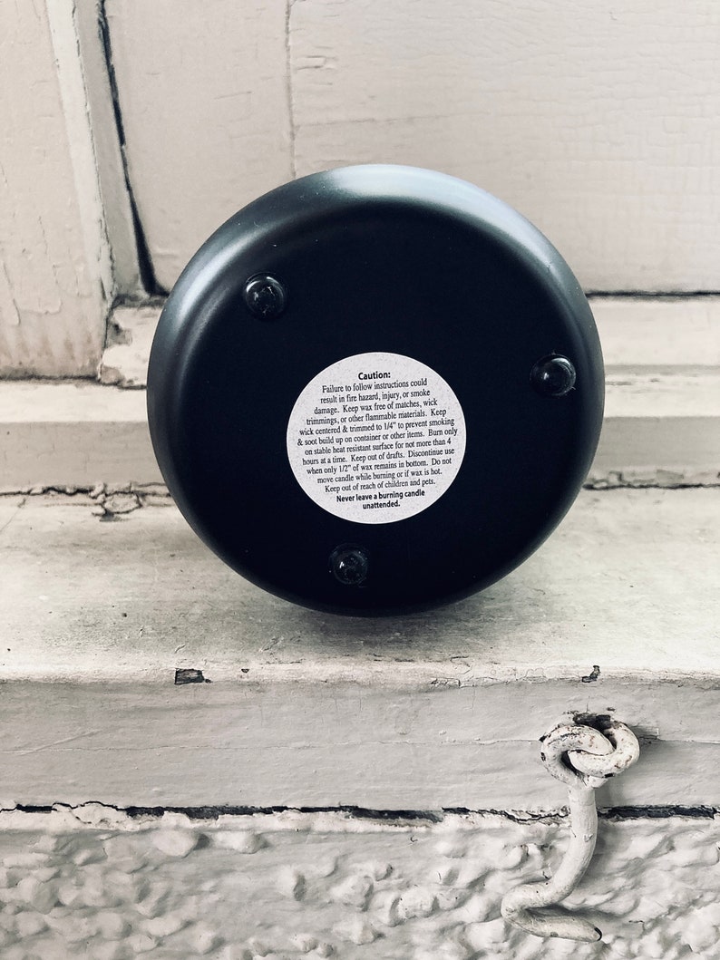 A black soy wax candle labeled “Cold Dead Heart” in gothic-style typography. It comes in a dark container with a matching lid featuring a minimalist triangle logo. The candle has a single white wick and a moody design perfect for gothic aesthetics.