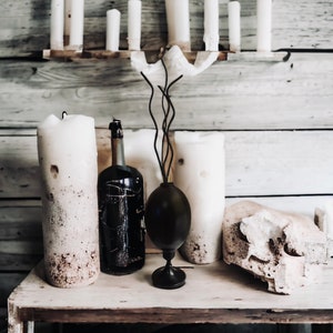 May include: A rustic still life featuring a black bottle, two white candles, a black vase with a twig-like design, and a white ceramic object on a wooden table.