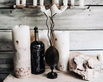 May include: A rustic still life featuring a black bottle, two white candles, a black vase with a twig-like design, and a white ceramic object on a wooden table.