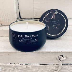 A black soy wax candle labeled “Cold Dead Heart” in gothic-style typography. It comes in a dark container with a matching lid featuring a minimalist triangle logo. The candle has a single white wick and a moody design perfect for gothic aesthetics.