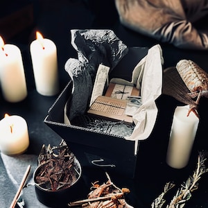 May include: A black box with a white tissue paper lining, filled with small, brown paper-wrapped items. The box is surrounded by candles and other decorative items.