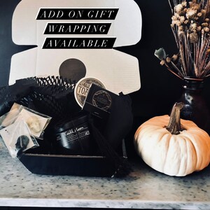 May include: A black gift box filled with various items, including a candle, a small tin, and a black pouch. The box is sitting on a marble surface next to a white pumpkin. The text "ADD ON GIFT WRAPPING AVAILABLE" is written on a white background.