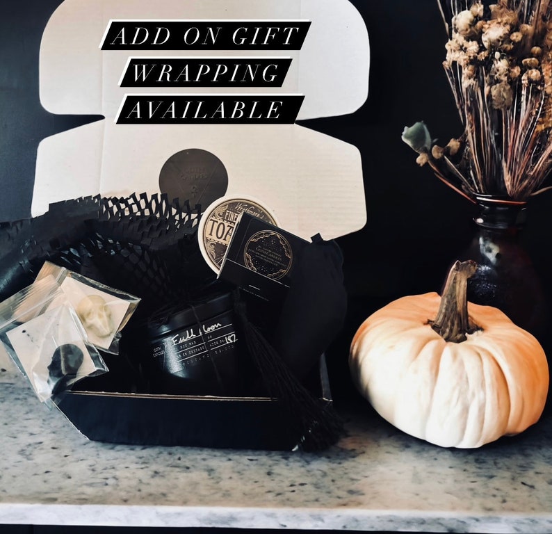 May include: A black gift box filled with various items, including a candle, a small tin, and a black pouch. The box is sitting on a marble surface next to a white pumpkin. The text "ADD ON GIFT WRAPPING AVAILABLE" is written on a white background.