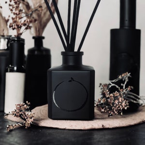 May include: Black diffuser bottle with black reeds and a circular design. The bottle is sitting on a light brown surface.
