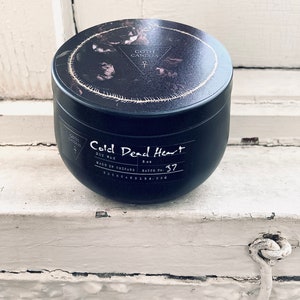 A black soy wax candle labeled “Cold Dead Heart” in gothic-style typography. It comes in a dark container with a matching lid featuring a minimalist triangle logo. The candle has a single white wick and a moody design perfect for gothic aesthetics.