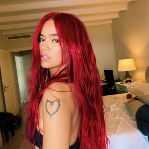 May include: A woman with long, bright red hair, wearing a black tank top and blue jeans. She has a small tattoo of a barbed wire heart on her left shoulder.