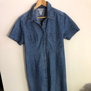 May include: A short-sleeved, button-down denim dress in a medium blue wash. The dress has a subtle leaf pattern and two chest pockets. It hangs on a wooden hanger against a white wall.