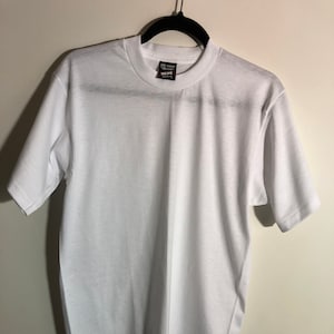 May include: A white t-shirt hanging on a black hanger. The t-shirt has a small tag on the front that says "Best" in black text.