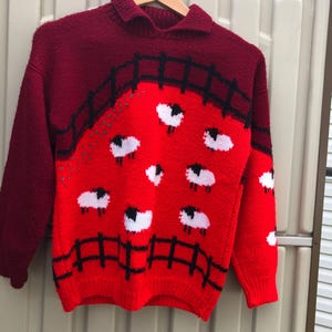 Peut inclure: Un pull en tricot rouge et bordeaux avec un motif de moutons noirs et blancs. Le pull a un col montant et des manches longues.