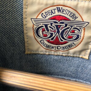 May include: Close-up of a light blue denim jacket with a tag. The tag reads "GREAT WESTERN GARMENT COMPANY" in a circular design with red and blue accents. The jacket is hanging on a wooden hanger.