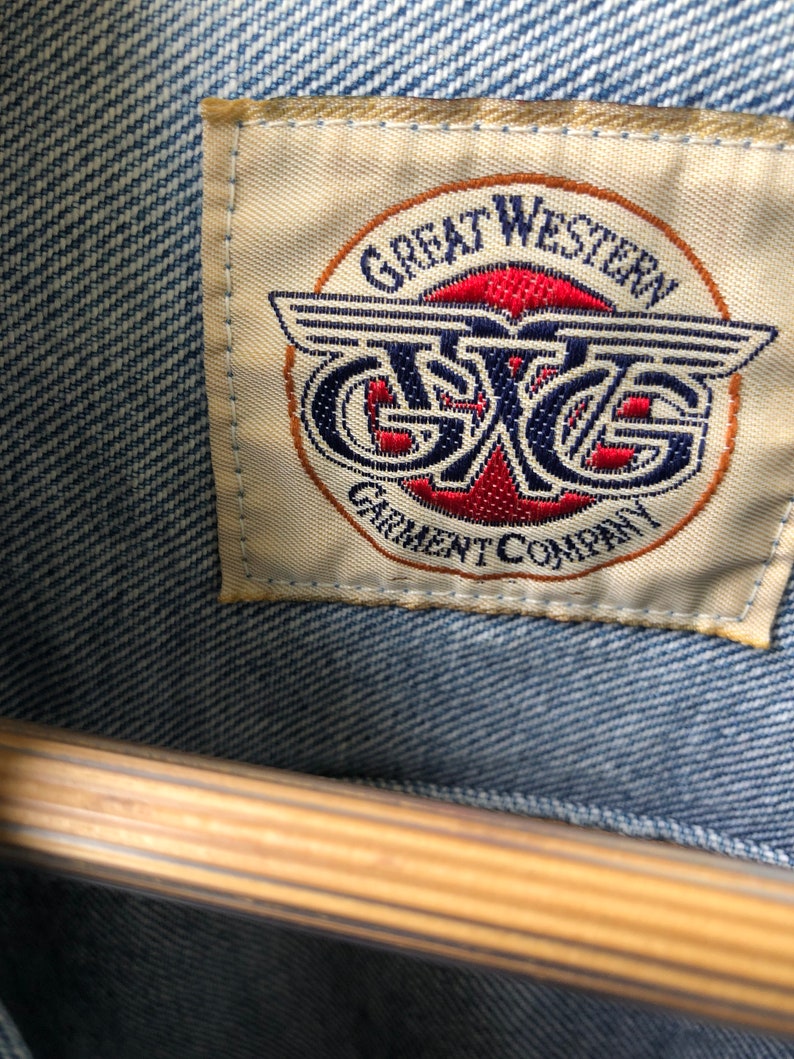 May include: Close-up of a light blue denim jacket with a tag. The tag reads "GREAT WESTERN GARMENT COMPANY" in a circular design with red and blue accents. The jacket is hanging on a wooden hanger.