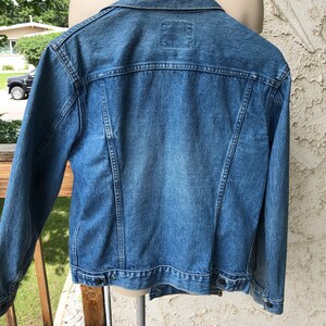 May include: A mid-wash denim jacket with a traditional design. The jacket has a collar, button fastenings, and a small square patch on the back. The jacket is displayed on a mannequin, showing its back and shoulder details. The jacket is hanging on a wooden hanger.