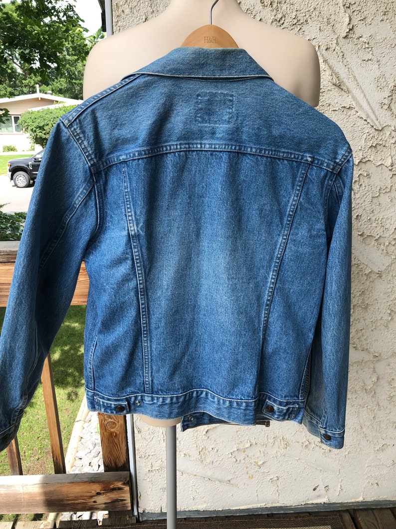 May include: A mid-wash denim jacket with a traditional design. The jacket has a collar, button fastenings, and a small square patch on the back. The jacket is displayed on a mannequin, showing its back and shoulder details. The jacket is hanging on a wooden hanger.