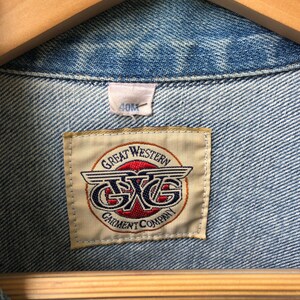 May include: Close-up of a light blue denim jacket with a visible label. The label reads "GREAT WESTERN GARMENT COMPANY" with a logo featuring the letters "GXG" in a circular design. The jacket is hanging on a wooden hanger.