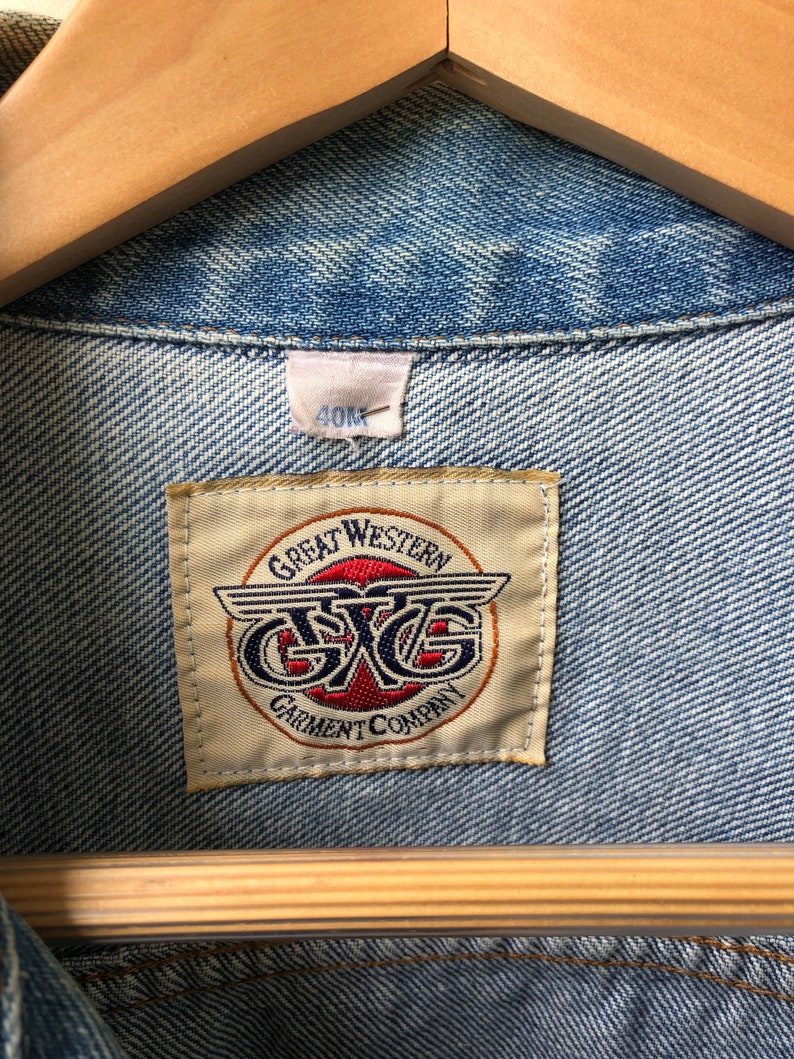 May include: Close-up of a light blue denim jacket with a visible label. The label reads "GREAT WESTERN GARMENT COMPANY" with a logo featuring the letters "GXG" in a circular design. The jacket is hanging on a wooden hanger.