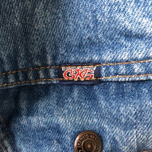 May include: Close-up of a blue denim jacket. The jacket features a small rectangular tag with the letters "GKS" in red and white. A bronze button with the words "WESTERN GARMENT GREAT" is also visible.