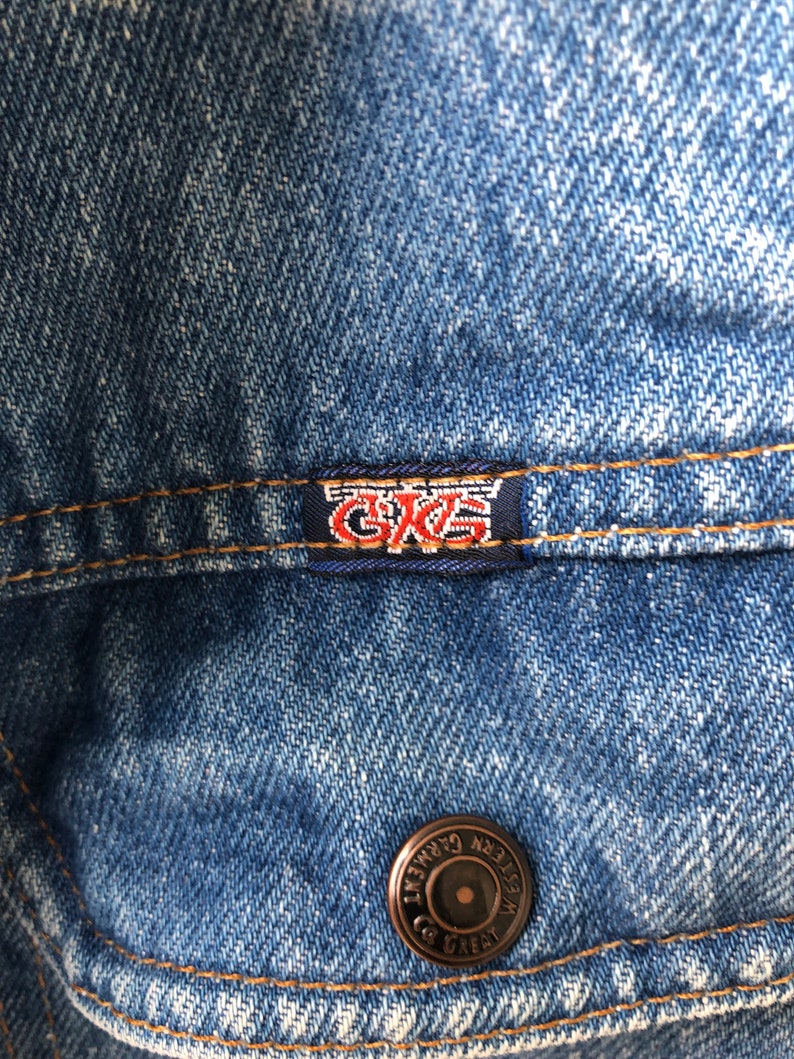 May include: Close-up of a blue denim jacket. The jacket features a small rectangular tag with the letters "GKS" in red and white. A bronze button with the words "WESTERN GARMENT GREAT" is also visible.