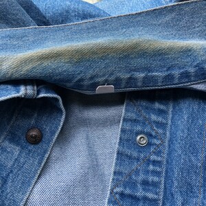 May include: Close-up of a blue denim jacket. The jacket has a button closure and a metal snap. The denim has a faded appearance with some discoloration. The stitching is visible, and the fabric is folded.